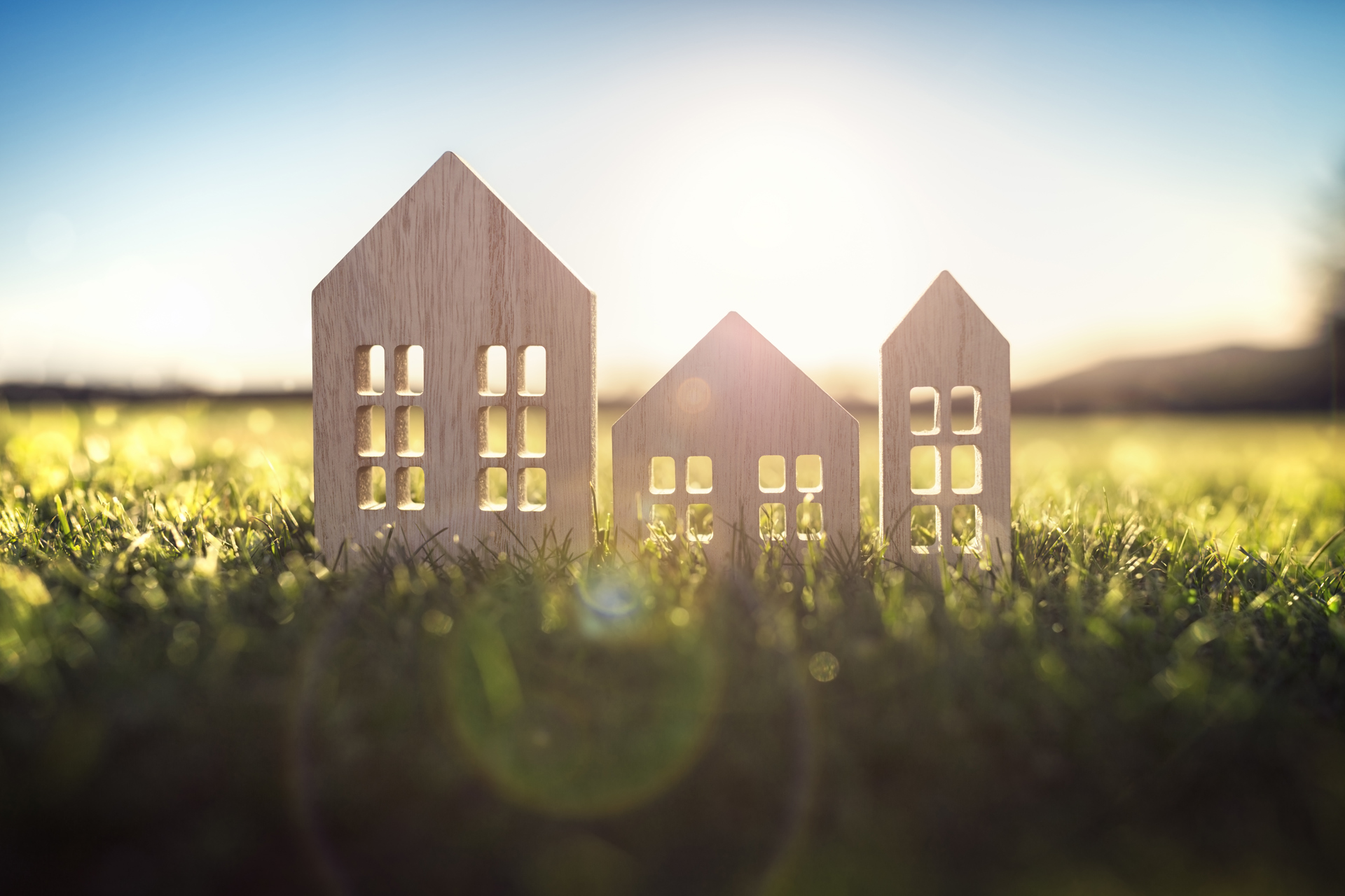 Sur l’herbe se trouvent trois découpes de bois plates en forme de maisons de styles différents, éclairées par le soleil. 